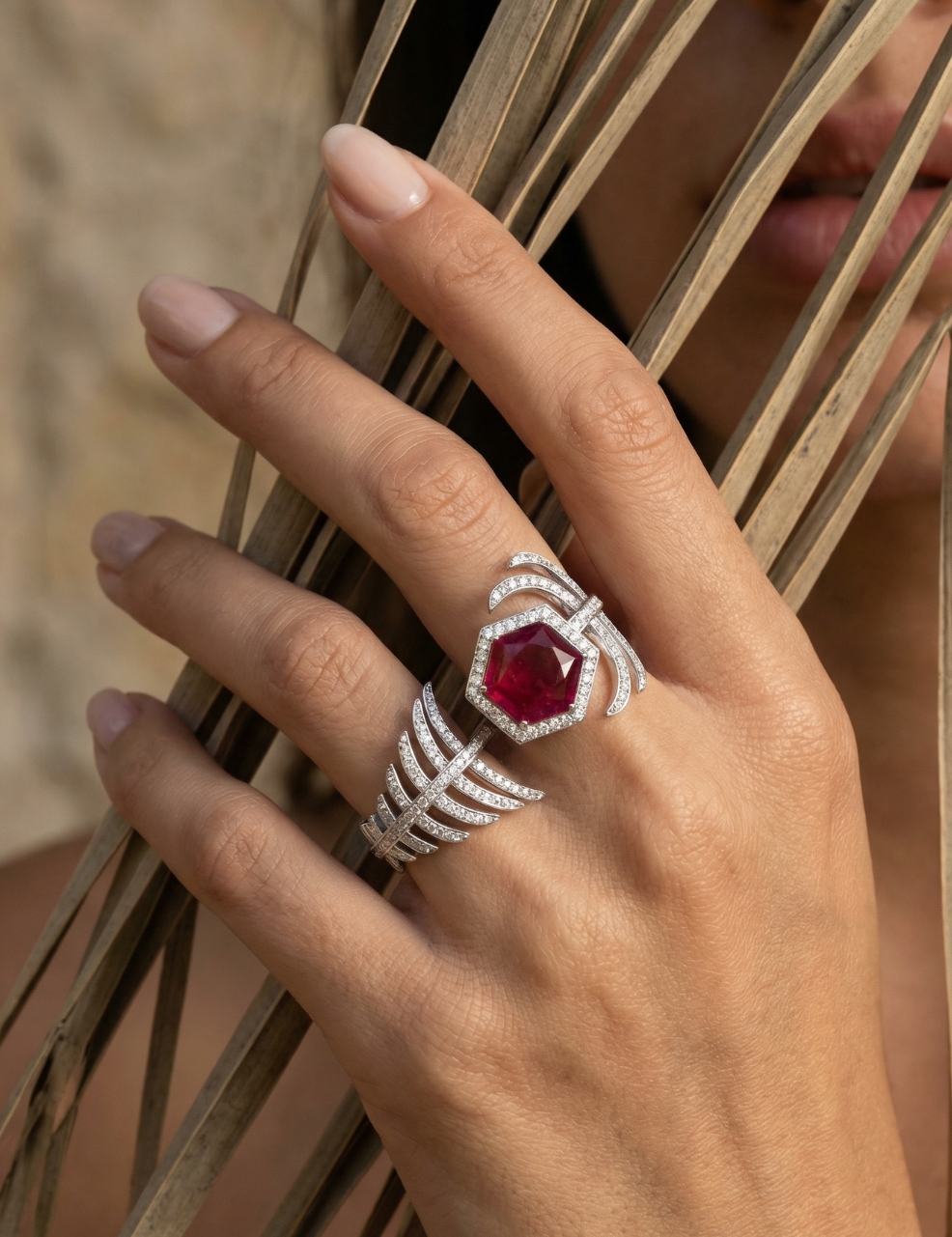 A close-up of a hand wearing a silver ring with a large hexagonal red gemstone and a matching smaller ring with curved silver bands and clear stones, set against dried palm leaves.