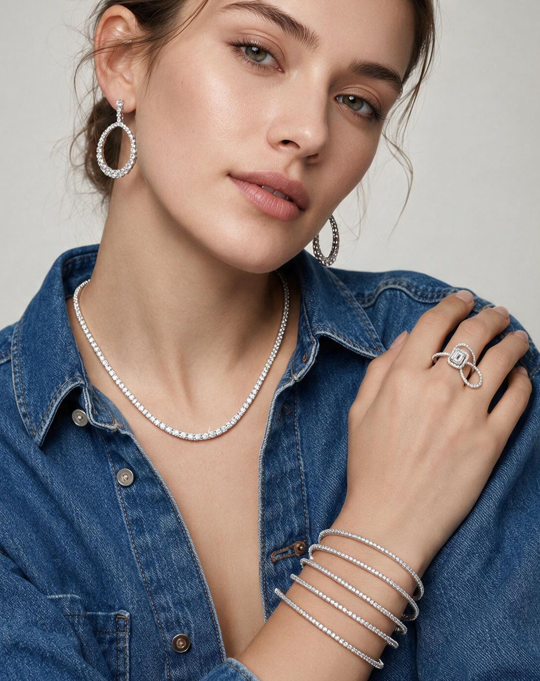 A close-up of a woman wearing a delicate diamond necklace, matching hoop earrings, and stacked diamond bracelets, styled with a blue denim shirt against a neutral gray background.