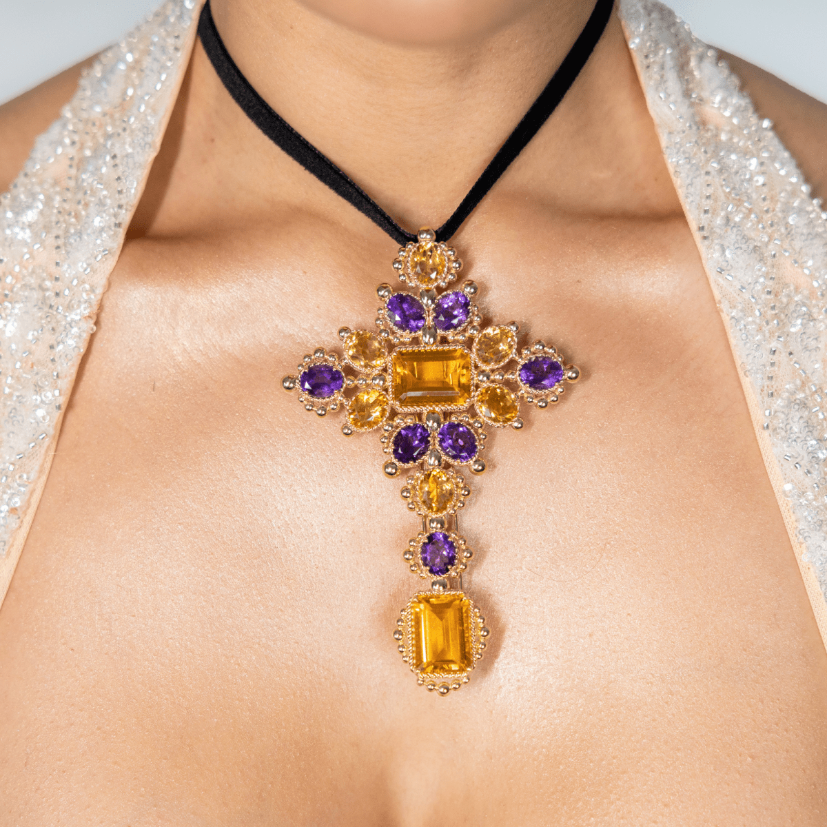 A close-up of a model wearing a gold cross pendant necklace with purple and yellow gemstones on a black cord, paired with a sparkling white halter top.