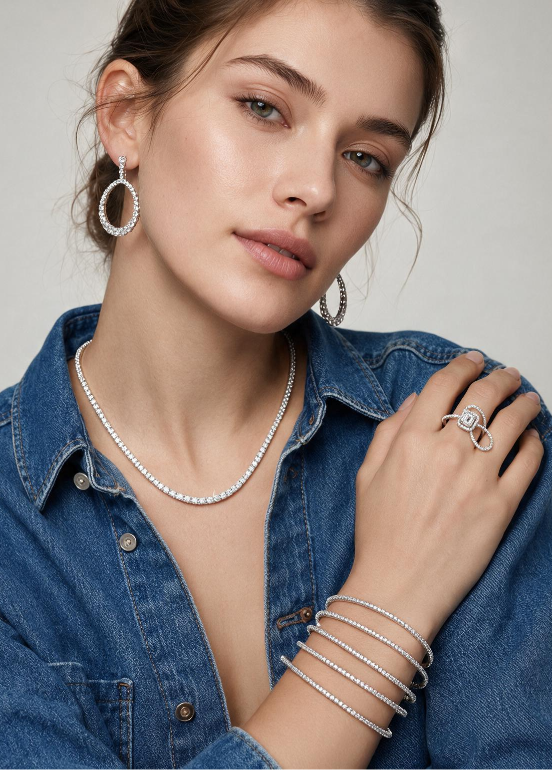 A close-up of a woman wearing a delicate diamond necklace, matching hoop earrings, and stacked diamond bracelets, styled with a blue denim shirt against a neutral gray background.