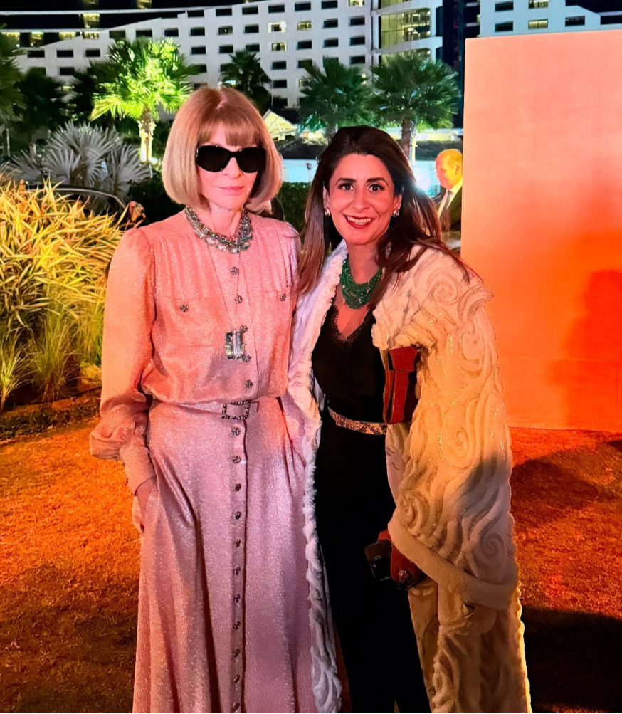 Two women posing together outdoors with a building and palm trees in the background wearing jewelry from Devi Jewels